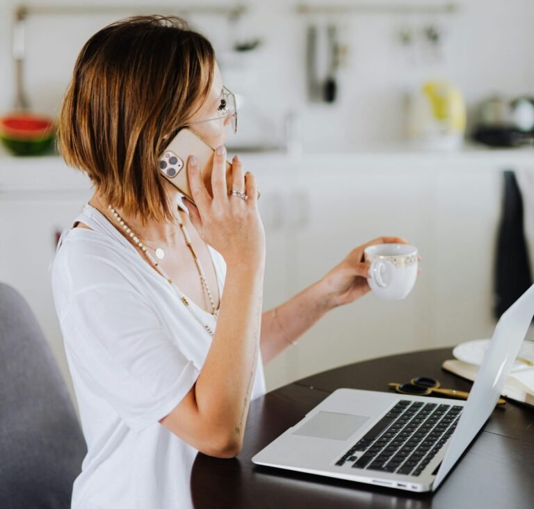 Woman multitasking with a smartphone and laptop while drinking coffee in a cozy home setting.